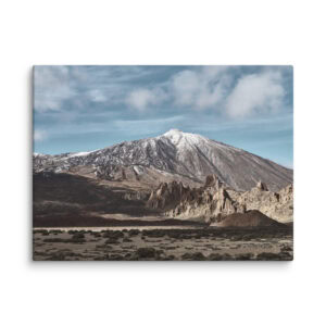 Una montaña nevada bajo un cielo azul nublado, acompañada de formaciones rocosas escarpadas y vegetación escasa en primer plano, captura la belleza natural de La Palma. Esta pintoresca escena se puede transformar en el "Lienzo del Teide", disponible exclusivamente a través de nuestra tienda online como arte decorativo.