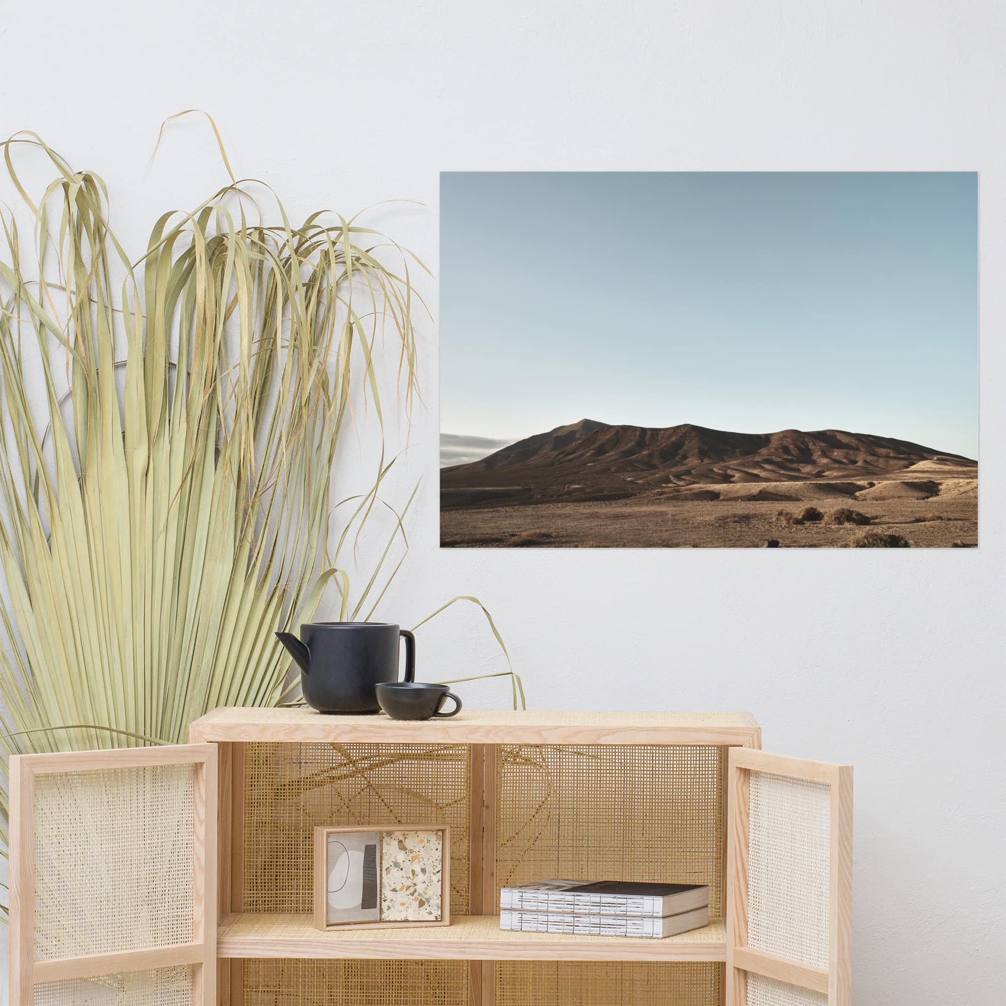 El interior minimalista cuenta con un armario de madera clara adornado con una tetera y una taza negras. A la izquierda, La Palma aporta elegancia, mientras que un póster enmarcado de Los Ajaches, que representa un paisaje desértico con colinas bajo un cielo azul claro, realza el ambiente sereno.