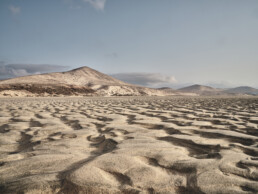 Un vasto paisaje desértico con onduladas dunas de arena bajo un cielo azul claro. Al fondo, varios picos de montañas se elevan majestuosos, creando una escena digna de un Póster Sotavento, que encarna el Arte Decorativo sereno y expansivo.