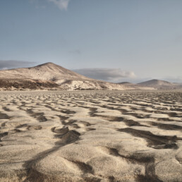 Un vasto paisaje desértico con onduladas dunas de arena bajo un cielo azul claro. Al fondo, varios picos de montañas se elevan majestuosos, creando una escena digna de un Póster Sotavento, que encarna el Arte Decorativo sereno y expansivo.