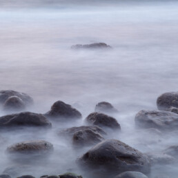 La imagen tranquila de rocas parcialmente sumergidas en el agua en La Palma, capturada con un efecto de exposición prolongada para crear una superficie brumosa, presenta una calidad etérea debido a las rocas dispersas de manera desigual y la iluminación suave. Es ideal para fotografías impresas o arte decorativo exquisitas, como el Póster de Rocas de la playa de El Lloret.
