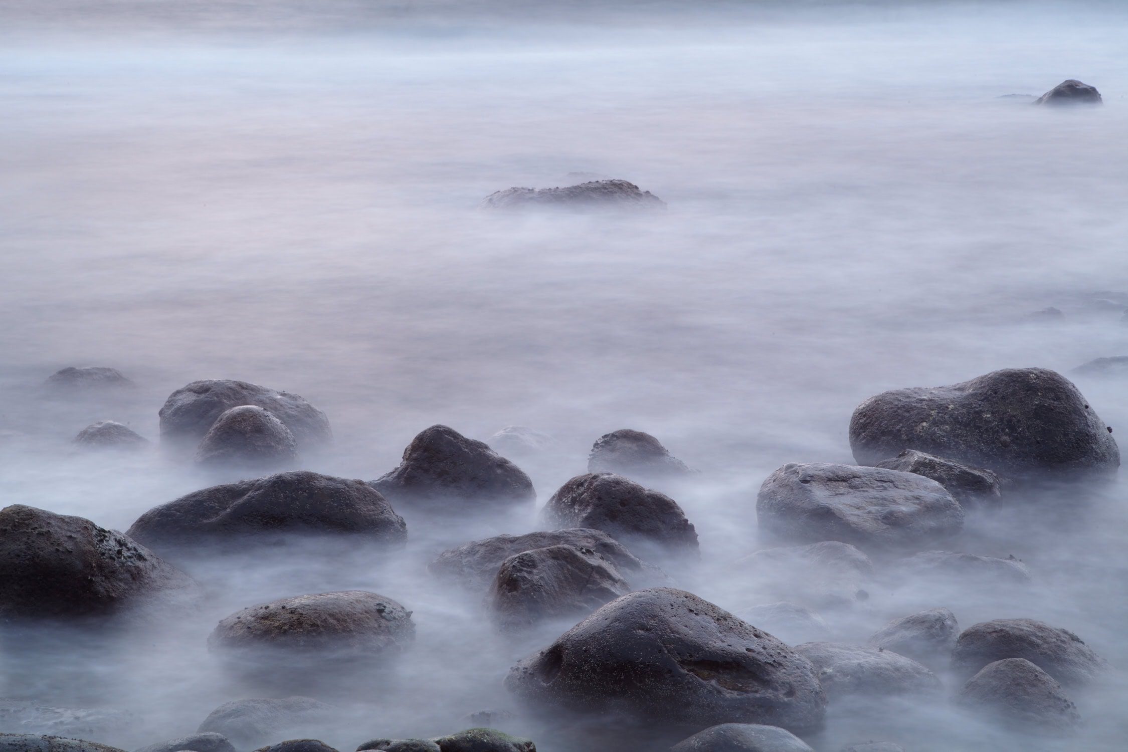 La imagen tranquila de rocas parcialmente sumergidas en el agua en La Palma, capturada con un efecto de exposición prolongada para crear una superficie brumosa, presenta una calidad etérea debido a las rocas dispersas de manera desigual y la iluminación suave. Es ideal para fotografías impresas o arte decorativo exquisitas, como el Póster de Rocas de la playa de El Lloret.