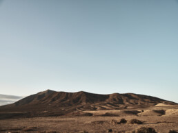 La esencia del Lienzo Los Ajaches se plasma en un vasto paisaje desértico con colinas áridas bajo un cielo azul claro. El terreno ondulado está marcado por una escasa vegetación y sombras del sol naciente o poniente, creando una vista serena y amplia.
