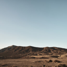 La esencia del Lienzo Los Ajaches se plasma en un vasto paisaje desértico con colinas áridas bajo un cielo azul claro. El terreno ondulado está marcado por una escasa vegetación y sombras del sol naciente o poniente, creando una vista serena y amplia.