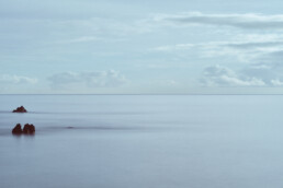Un tranquilo paisaje marino con suaves olas y un cielo azul suave captura la esencia de Paisajes Canarios. Formaciones rocosas sobresalen del agua en primer plano, mientras nubes esponjosas salpican el horizonte. Esta vista serena es perfecta para que Arte Decorativo o Fotografías impresas evoquen calma.