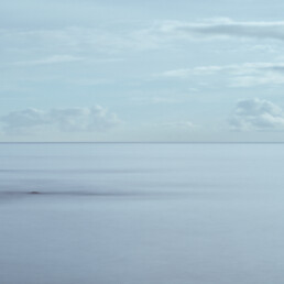 Un tranquilo paisaje marino con suaves olas y un cielo azul suave captura la esencia de Paisajes Canarios. Formaciones rocosas sobresalen del agua en primer plano, mientras nubes esponjosas salpican el horizonte. Esta vista serena es perfecta para que Arte Decorativo o Fotografías impresas evoquen calma.