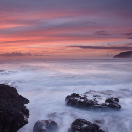 Una tranquila vista costera de Los Guirres al atardecer muestra una costa rocosa con olas que acarician suavemente las rocas. El cielo está adornado con tonos de rosa, naranja y violeta, con acantilados recortados en el horizonte, una captura ideal para Póster rocas de Los Guirres como una exquisita pieza de arte decorativo.
