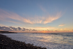 Atardecer en una playa rocosa de La Palma, donde las olas acarician suavemente la orilla y el cielo se tiñe de tonos naranjas, rosas y azules. Las nubes dispersas realzan la escena y una silueta de tierra lejana adorna el horizonte, una representación perfecta capturada en 