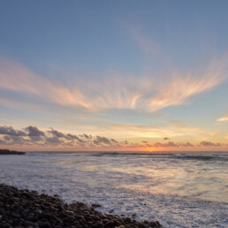 Atardecer en una playa rocosa de La Palma, donde las olas acarician suavemente la orilla y el cielo se tiñe de tonos naranjas, rosas y azules. Las nubes dispersas realzan la escena y una silueta de tierra lejana adorna el horizonte, una representación perfecta capturada en 