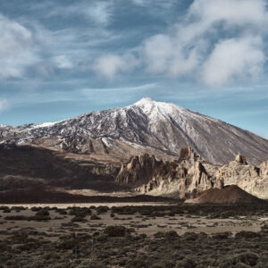 Una vista panorámica de una montaña cubierta de nieve bajo un cielo parcialmente nublado muestra el arte de la naturaleza que recuerda a una obra maestra del arte decorativo. En primer plano se ven afloramientos rocosos y un paisaje desértico, donde las escarpadas laderas de la montaña contrastan maravillosamente con el terreno extenso y llano.