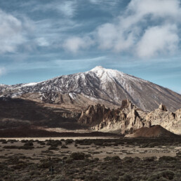 Una vista panorámica de una montaña cubierta de nieve bajo un cielo parcialmente nublado muestra el arte de la naturaleza que recuerda a una obra maestra del arte decorativo. En primer plano se ven afloramientos rocosos y un paisaje desértico, donde las escarpadas laderas de la montaña contrastan maravillosamente con el terreno extenso y llano.