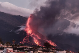 Un volcán entra en erupción con lava de un rojo intenso y humo negro denso, sobre el fondo montañoso y nublado de La Palma. En primer plano, hay un pueblo con casas y palmeras, que ofrece un contraste sorprendente con el espectáculo volcánico ardiente capturado en el 