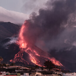 Un volcán entra en erupción con lava de un rojo intenso y humo negro denso, sobre el fondo montañoso y nublado de La Palma. En primer plano, hay un pueblo con casas y palmeras, que ofrece un contraste sorprendente con el espectáculo volcánico ardiente capturado en el 
