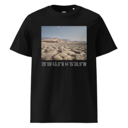 La camiseta unisex Sotavento de nuestra tienda online es una camiseta negra adornada con una imagen de un paisaje desértico con dunas de arena y montañas lejanas. Debajo de esta impresionante imagen, las coordenadas 28°0643.0N 14°1538.9W están impresas en texto blanco, capturando la esencia de las cautivadoras vistas de La Palma.