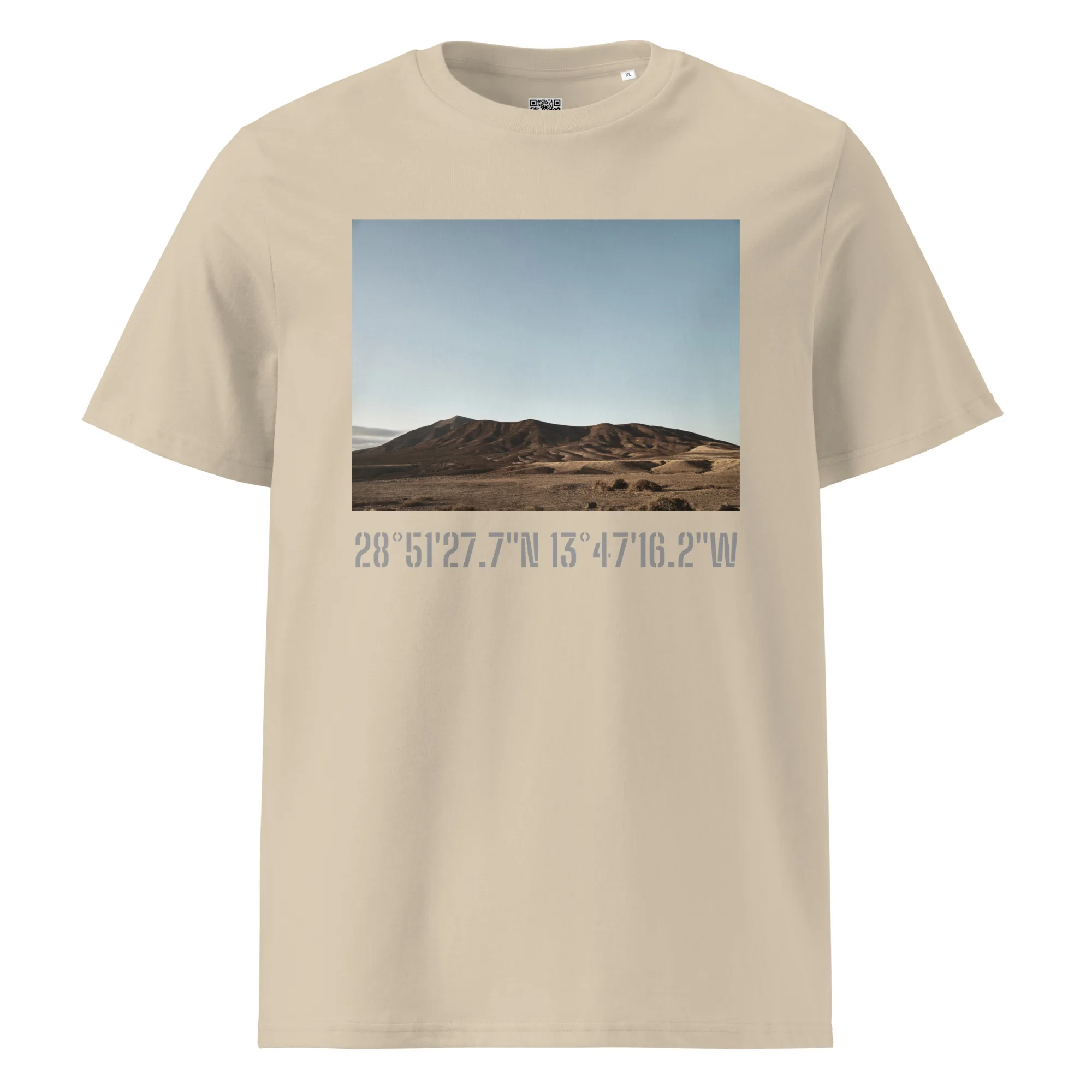 Camiseta unisex Los Ajaches en color beige, con una impresionante fotografía impresa de un paisaje desértico que muestra colinas onduladas bajo un cielo azul claro. Debajo de la imagen se encuentran impresas las coordenadas: 28°51'27.7"N 13°47'16.2"W, capturando la esencia de los paisajes canarios.