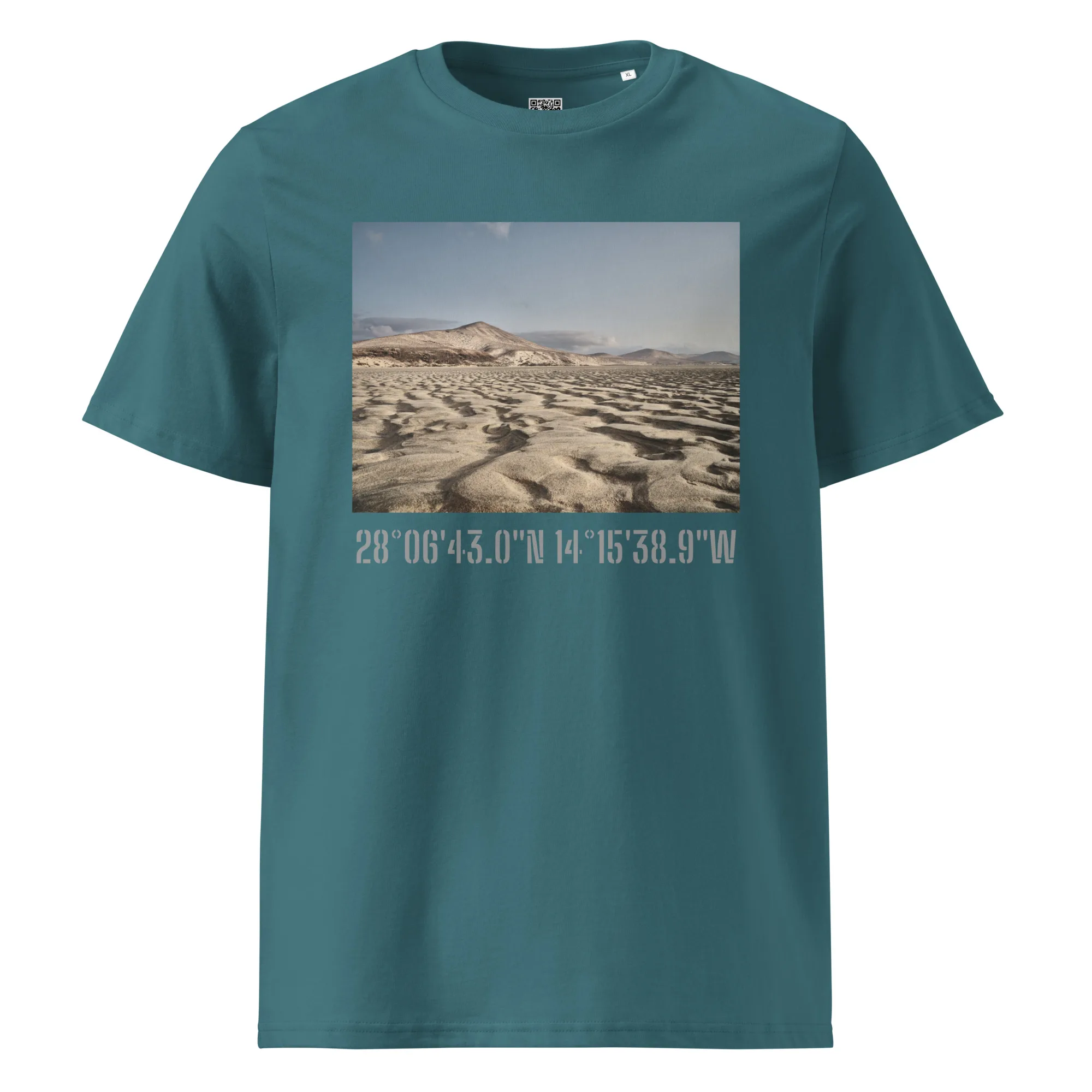 La camiseta unisex Sotavento, diseñada por Volcanica Productions y vendida exclusivamente en Tienda Online, presenta un color verde azulado con una fotografía cautivadora de un paisaje desértico con dunas y montañas bajo un cielo azul claro. El diseño está inspirado en el espectacular terreno cerca de La Palma, con las coordenadas 28°0643.0N 14°1538.9W que se muestran debajo de la imagen.